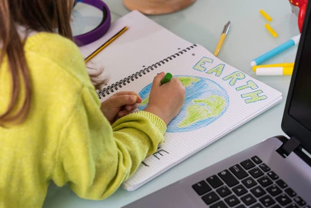 A child wearing a yellow sweater is drawing the planet Earth on a notebook. The word “Earth” is colored in green.