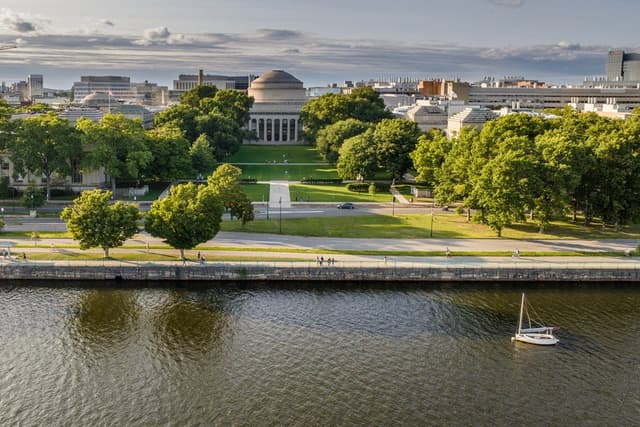 Aerial photo of MIT’s Great Dome with a lawn, trees, and a river in the foreground.